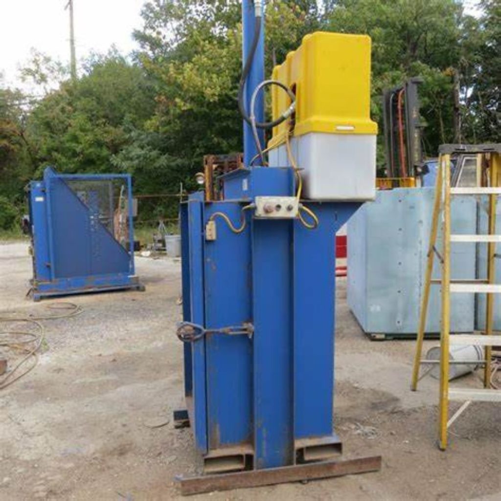 A blue industrial hydraulic baler, marked for the Department: Mining and Gas, stands outdoors on a concrete surface. Tall and rectangular with a yellow control box on top, it is surrounded by other industrial equipment pieces and trees in the background.