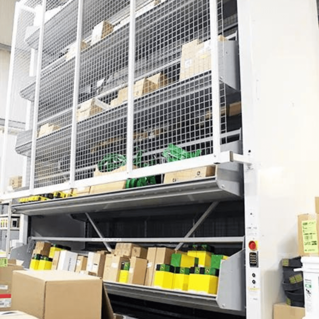 A vertical storage system with multiple shelves holds various-sized boxes, ideal for material handling in a warehouse. The white structure features metal mesh doors and a side control panel, reminiscent of a gallery exhibit. Some boxes are stacked on the floor ahead.