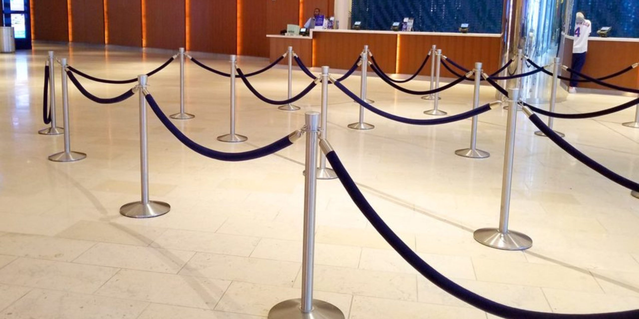 The empty hotel lobby, adorned with a tiled floor and Visiontron Products stanchions forming a zigzag queue, exudes warmth. A reception desk stands in the background under gentle lighting, offering seamless crowd control solutions.