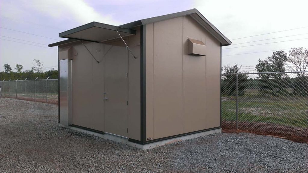 A small, beige utility building with a sloped roof and a metal door serves as a secure equipment storage, surrounded by a chain-link fence on gravel ground. The sky is overcast, and trees are visible in the background.