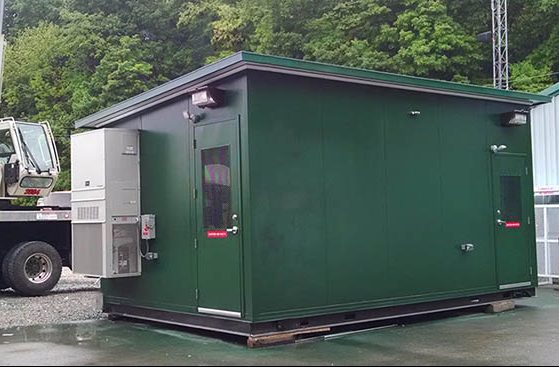 A green industrial container, serving as a modular equipment enclosure with air conditioning units, is situated on a concrete surface. In the background, a crane looms near a forested area, showcasing advanced equipment protection solutions.