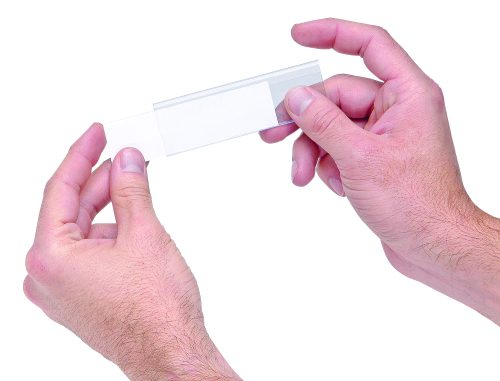 LTR-0813.jpg Two hands hold a clear microscope slide against a white background, fingers grasping gently on either side. It demonstrates precision akin to Quantum Label Holders with laser label inserts (25 per pack), underscoring meticulous attention to detail.