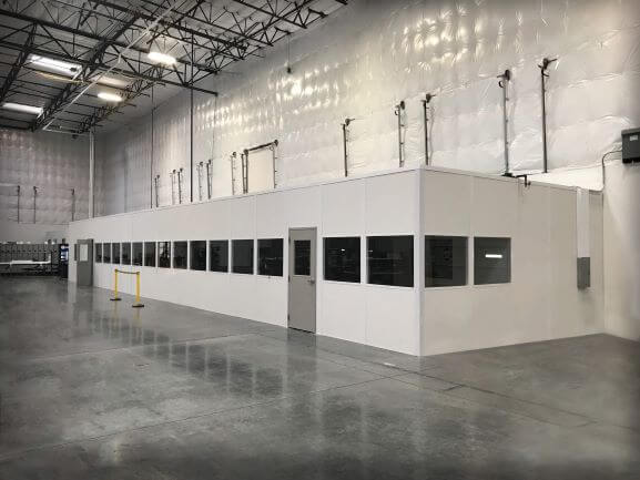 A large, white modular cleanroom with several windows and a door sits in a spacious industrial facility. Designed as an Industrial Equipment Enclosure, it boasts high ceilings and is surrounded by concrete flooring, while fluorescent lights illuminate the pristine workspace.
