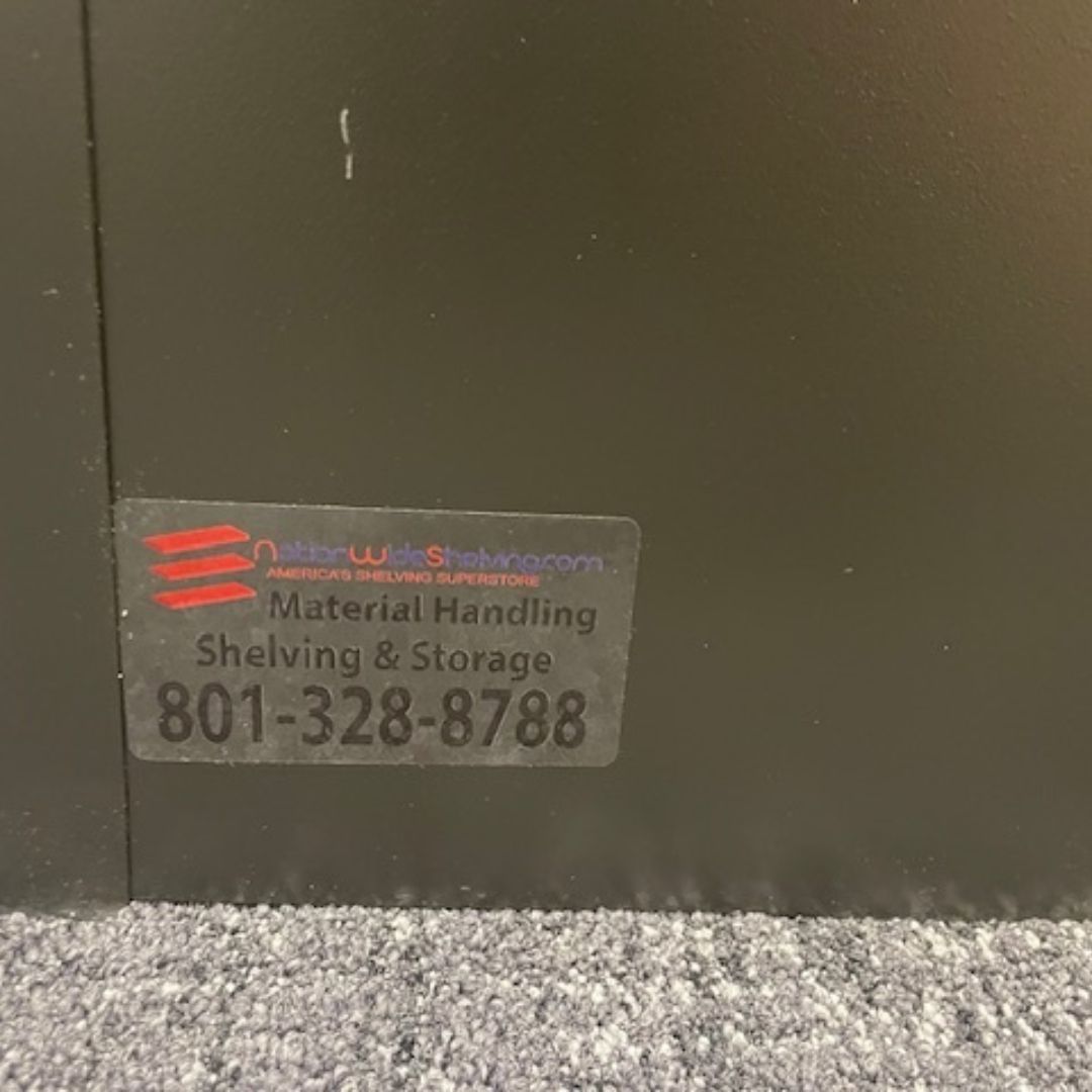 A black cabinet sports a sticker stating: Material Handling Shelving & Storage alongside the phone number 801-328-8788. Its durable design fits perfectly atop gray carpet, offering modular shelving solutions for financial institutions.