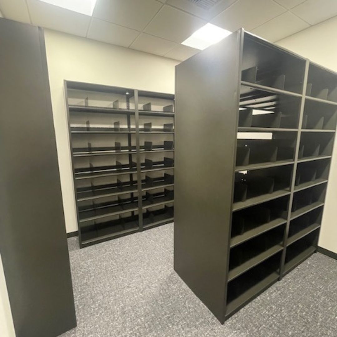 The room features empty gray metal shelving units, perfect for 40-Year Mortgage File Storage, arranged along the walls and center. With uniform divisions, the shelves rest on a carpeted floor under a white ceiling with fluorescent lighting.