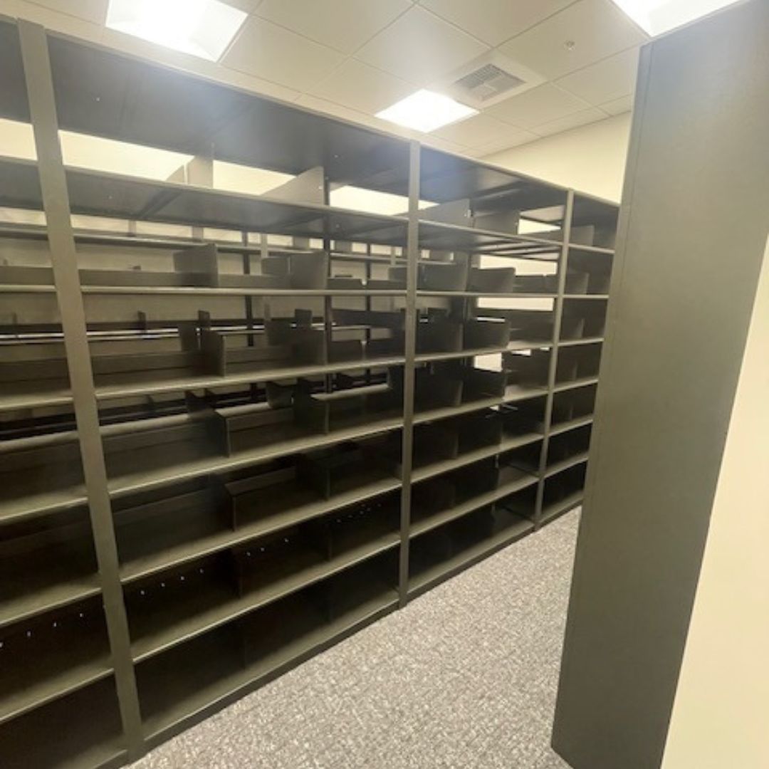 The empty dark wooden shelves, designed as modular shelving solutions for financial institutions, stretch from the foreground to the background in a brightly lit room with a carpeted floor.