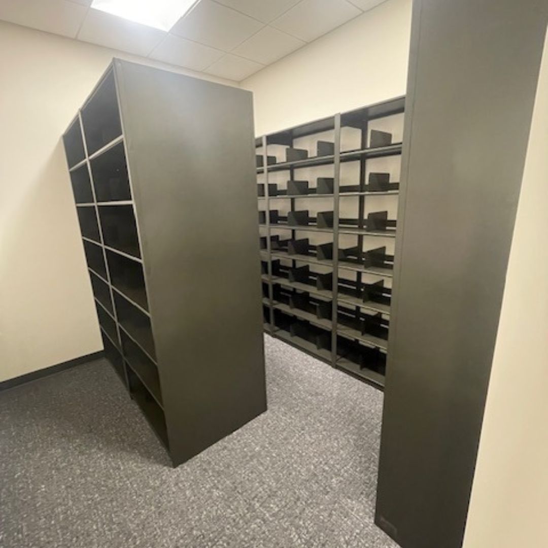 The storage room, designed for Mountain America Credit Union files, features dark metal shelving units on light gray carpet. Aurora Quik-Lok Shelving forms aisles and wall rows under recessed lighting in a pristine white ceiling, perfectly suited for organized 40-year mortgage file storage.