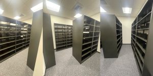 Panoramic view of an empty room with Aurora Quik-Lok Shelving lining the walls and center under fluorescent lighting. The durable, adjustable shelving systems are tall and spacious, while the floor is covered with gray carpet.