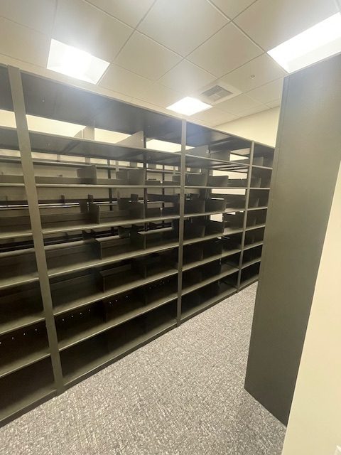 The room features rows of empty, dark-colored metal shelves, durable and adjustable for varied needs, under a fluorescent ceiling reflecting bright lights above. Carpeted floors enhance the readiness of this Mountain America Credit Union Storage space for 40-year mortgage file storage.