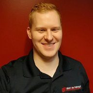 A person with short, red hair smiles while wearing a black collared shirt with red and white text. The solid red wall in the background subtly complements the organized vibe of athletic storage solutions.