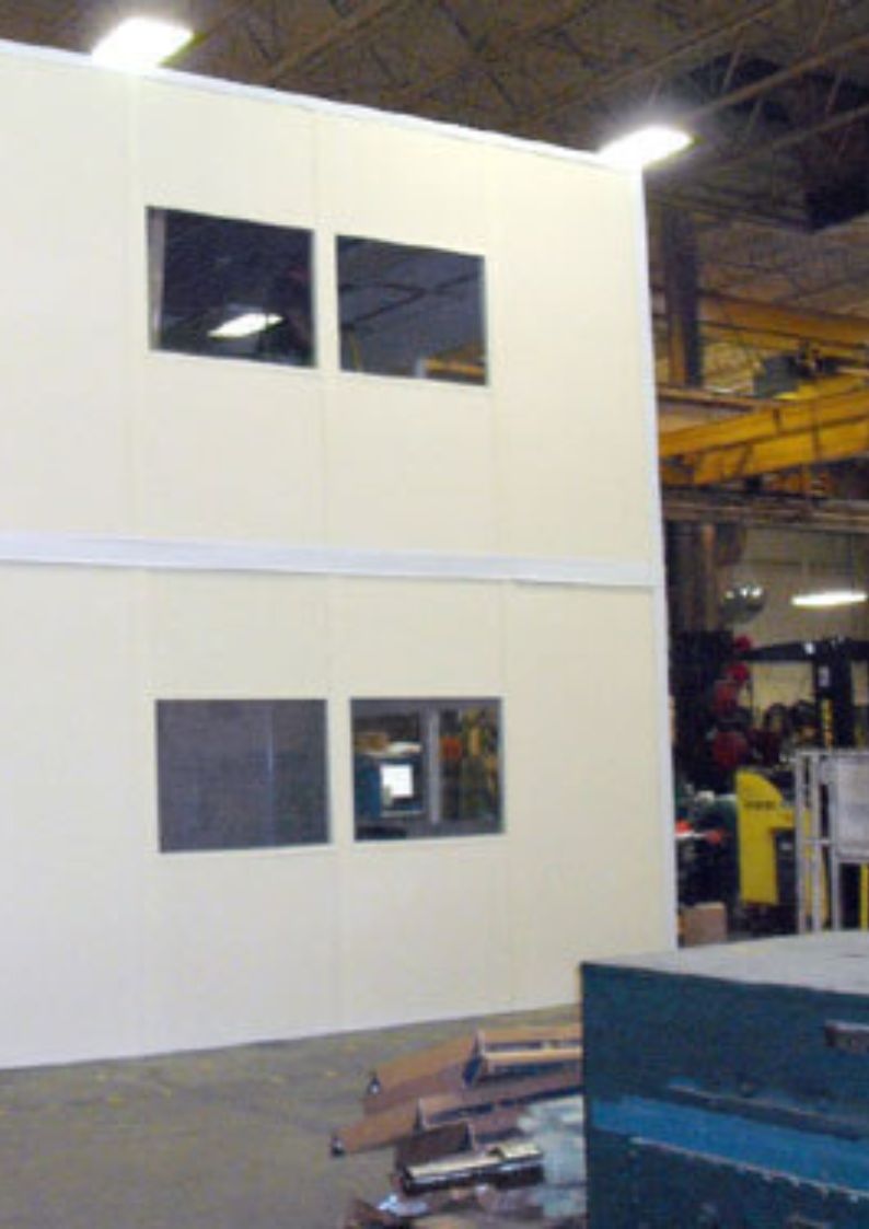 A large warehouse interior features a two-story, modular building with four rectangular windows. The space contains industrial equipment, including a yellow machine and various tools in the background. The ceiling is visible with metal beams and lighting.