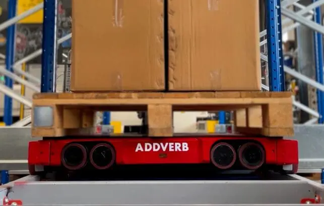 Red 4-way pallet shuttle transporting a wooden pallet with cardboard boxes inside a warehouse racking system.