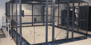 A large, secure metal cage enclosure with a locked gate, designed for optimal cage security, located indoors—possibly in a server or data center room with visible equipment and cables outside the cage.
