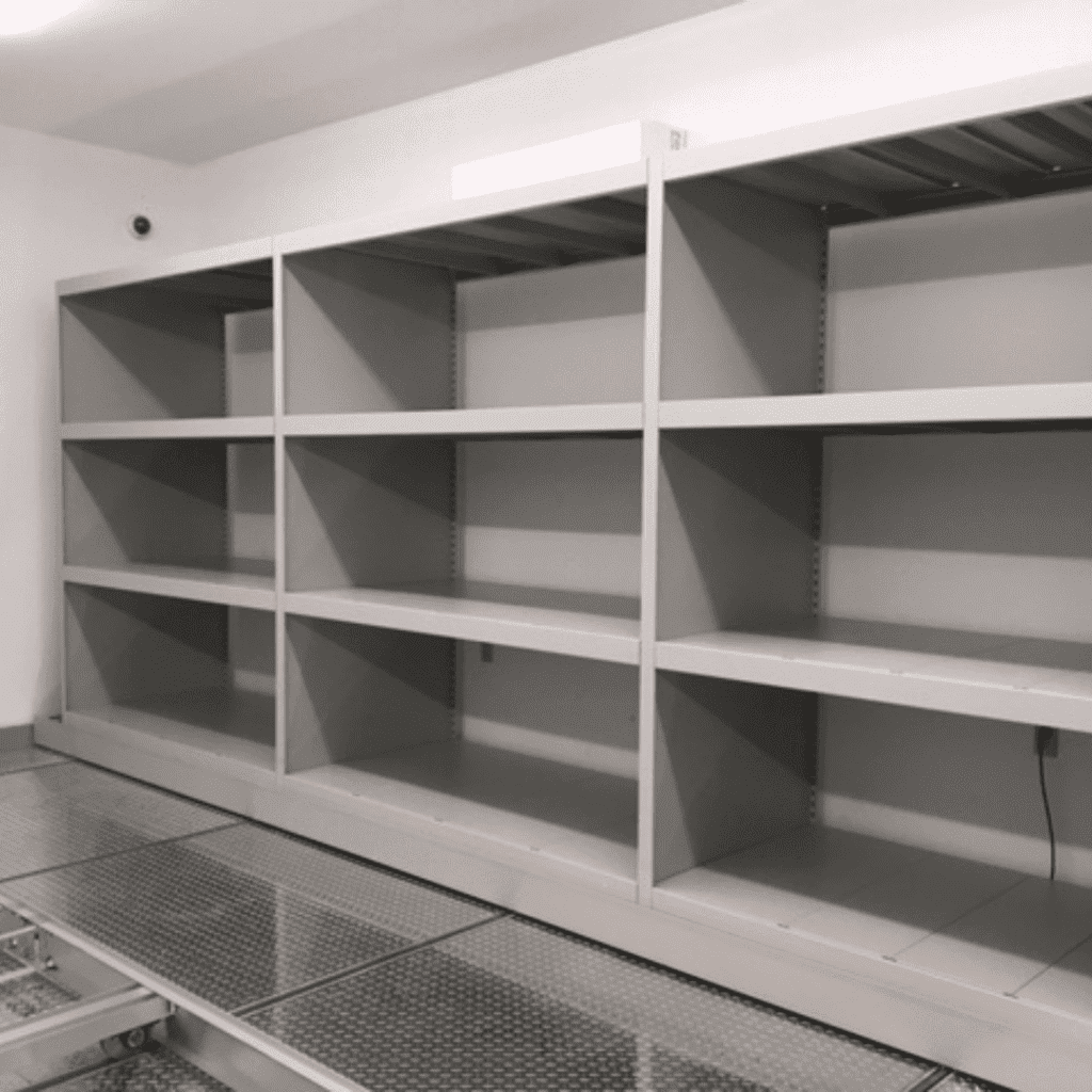 Large, empty metal shelves line a wall in a room with tiled flooring and bright lighting. The shelves have no items on them, creating a sparse and orderly appearance.