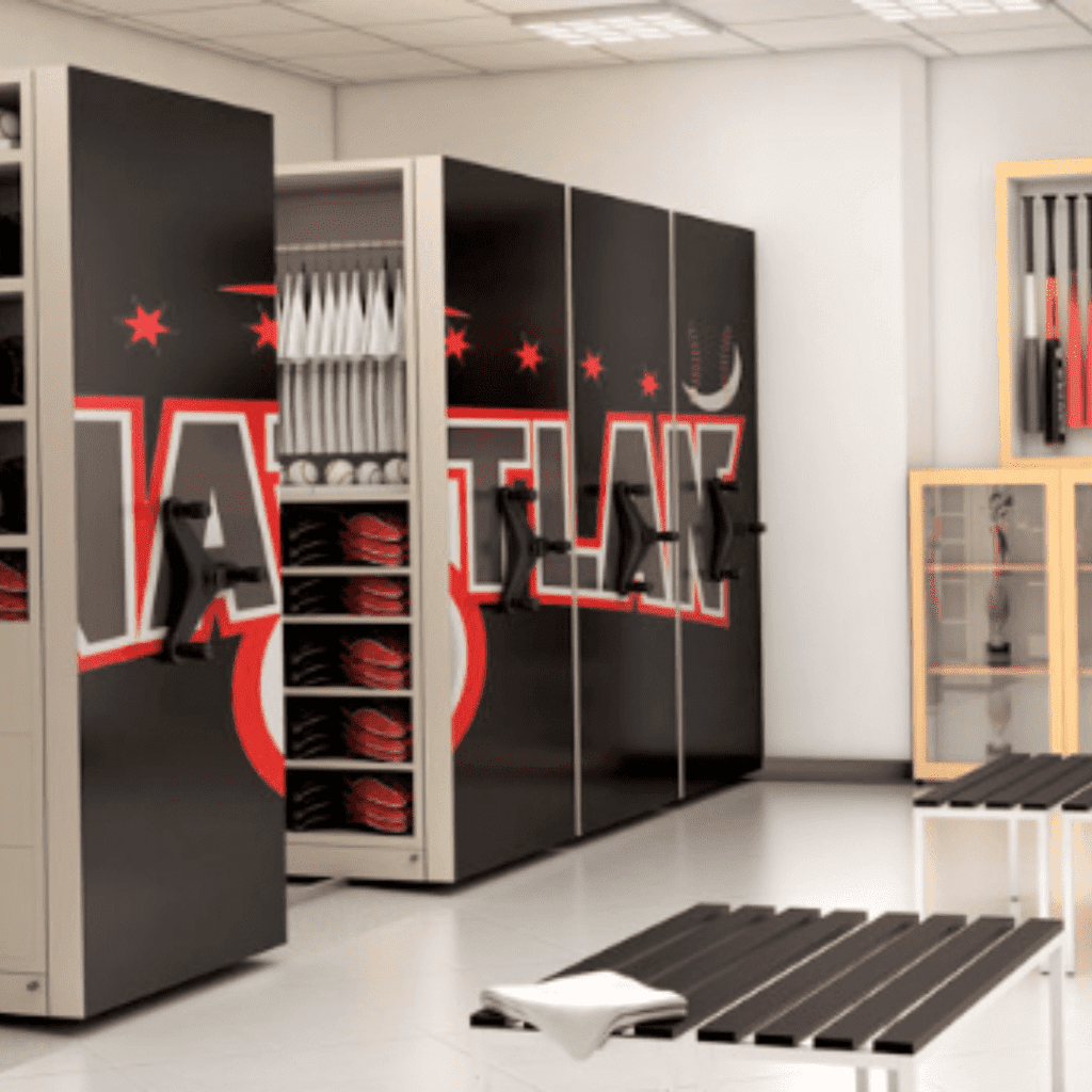 A modern locker room with black and red storage units holding helmets and bats, benches with folded towels, and a display case containing more sports equipment. The room is brightly lit and organized.