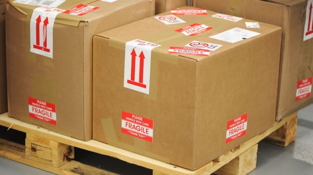 Several cardboard boxes labeled “Fragile” and “This Side Up” are stacked on wooden pallets, featuring frequently purchased shipping & packing supplies, ready for shipping or storage in a warehouse or shipping area.