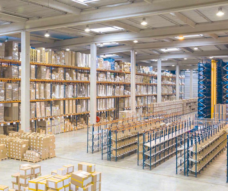 Spacious warehouse with high shelves filled with stacked cardboard boxes and goods, partially empty blue metal racks, and additional boxes organized on the floor. Bright lighting enhances this ergonomic warehouse layout for forklift and pedestrian safety.
