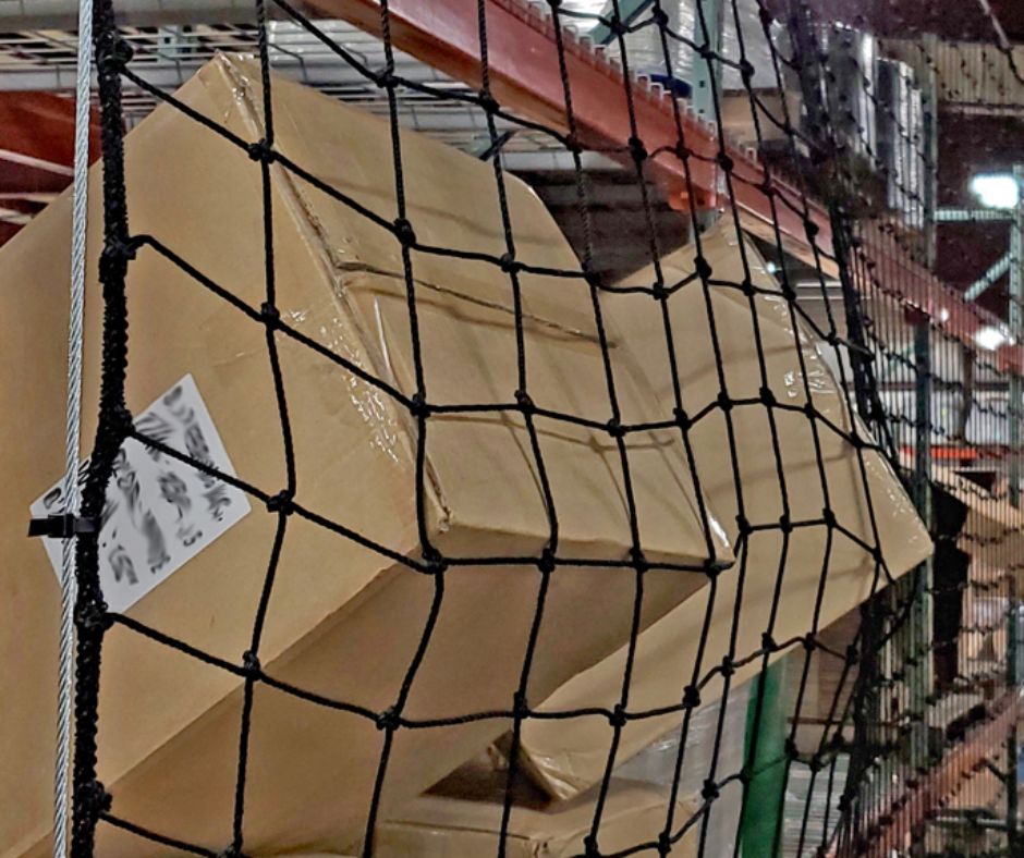 Large, damaged cardboard boxes are stacked on metal shelving with pallet rack backing and secured behind a black safety net in a warehouse. One box has a shipping label and appears partially crushed.