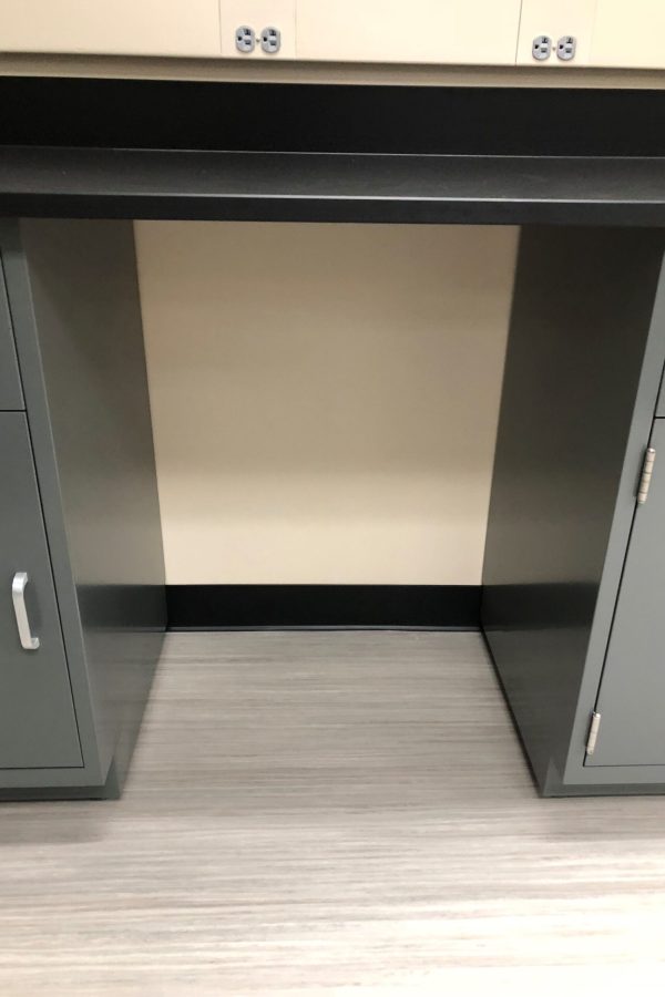 A gray desk with two cabinets and metal handles stands proudly against a beige wall, reminiscent of the studious ambiance found in a Weber State University dorm. The light wood floor complements the setting, while electrical outlets are strategically placed at the top on the wall.