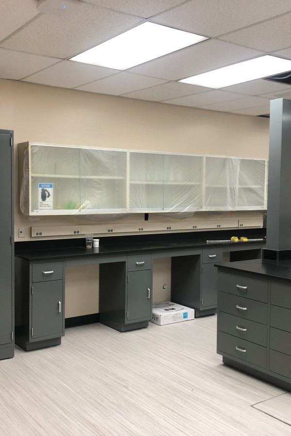 A modern laboratory at Weber State University features gray cabinets and countertops. Shelves are covered with protective plastic, ceiling tiles are partially missing, and a cardboard box rests on the floor. The room is well-lit with fluorescent lighting, maintaining an efficient workspace.